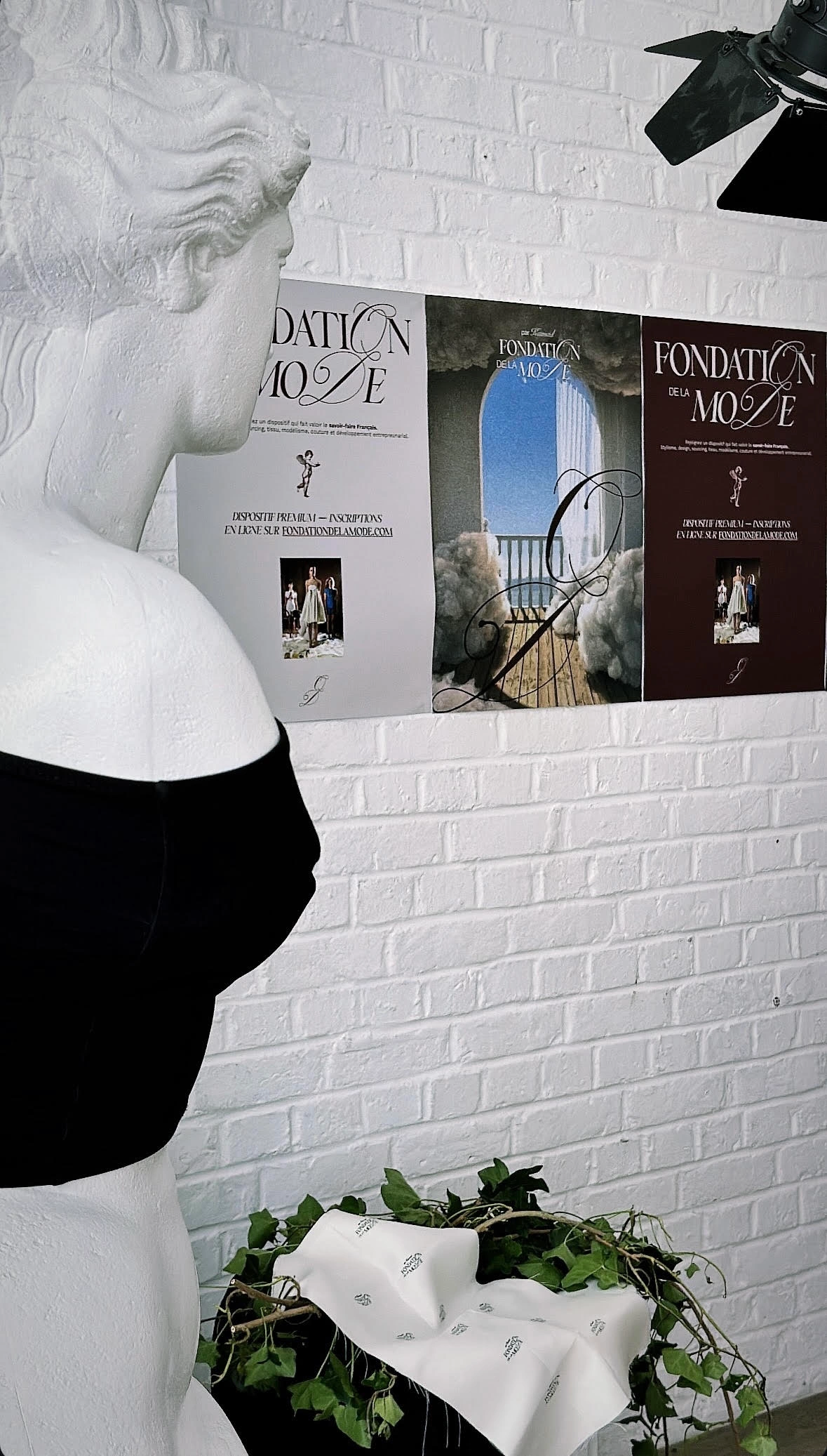 Affiches de la Fondation de la Mode sur un mur blanc, près d’un buste.